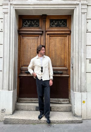 Camisa de botones color beige claro, jeans negros y zapatos oscuros. De pie junto a una puerta de madera con detalles ornamentales, con escalones de piedra plana abajo.