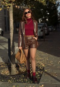 Blazer de cuero marrón sobre un suéter de cuello alto burdeos, acompañado de pantalones cortos de cuero marrón, bolso marrón y zapatos oscuros con calcetines rojos.