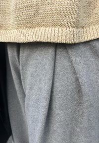 Bézs színű kötött pulóver alsó része szürke pliszírozott nadrág felett, amely texturált mintát és puha anyagot mutat. A színek kontrasztja jelentős.