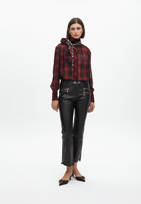 Blusa a scacchi rossa e nera con colletto annodato, abbinata a pantaloni in pelle nera con zip dorate e orli a spacco. Tacchi neri con dettagli.