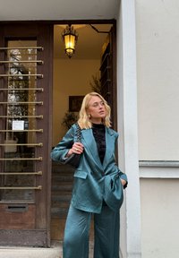 Grønnblå satengdress med dobbelspent blazer og vide bukser, kombinert med en svart blonder-topp og en teksturert svart håndveske.