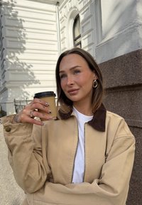 Giacca in coda di topo beige con colletto marrone, maglietta bianca sotto, tiene in mano un bicchiere di carta marrone per caffè, con unghie decorate e gioielli d'argento.