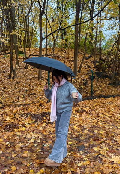 Paraguas negro sostenido por encima. Suéter gris de punto, bufanda rosa claro, pantalones acampanados grises y botas color café claro. Hojas de otoño cubren el suelo.