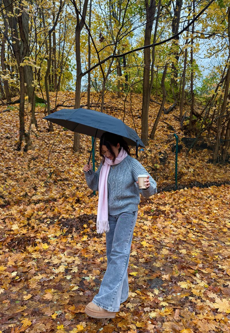 Guarda-chuva preto segurado por cima da cabeça. Suéter cinza de malha, cachecol rosa claro, calças cinza largas e botas castanho-claro. Folhas de outono cobrem o chão.