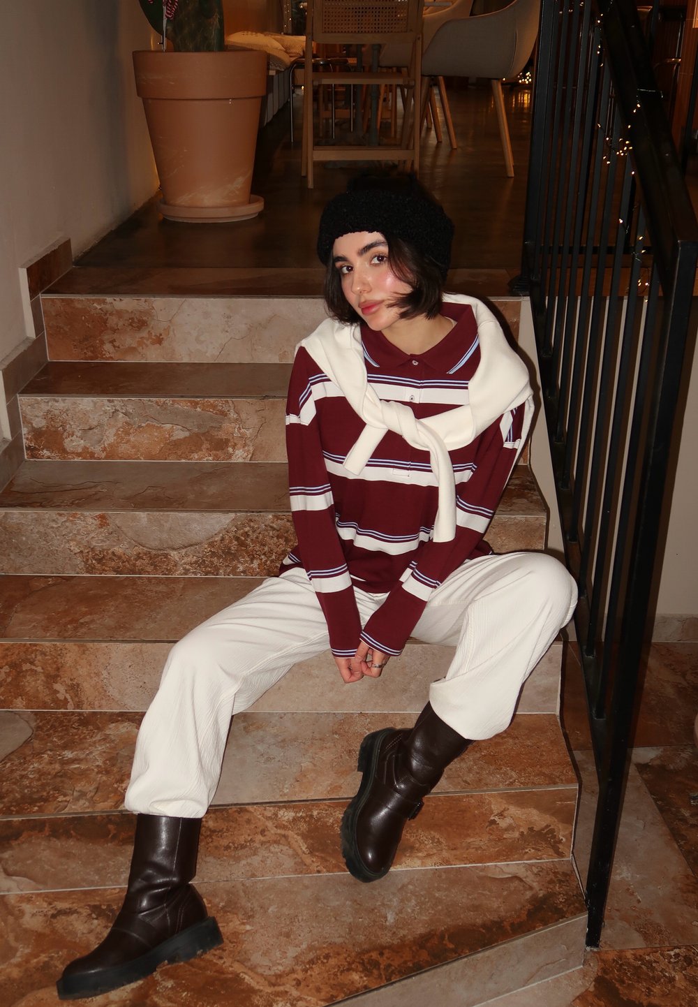 Maglietta a maniche lunghe a righe bordeaux e bianche con un maglione bianco appoggiato sulle spalle, abbinata a pantaloni crema e stivali marroni fino al ginocchio.