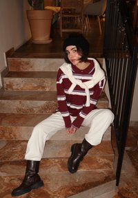 Maglietta a maniche lunghe a righe bordeaux e bianche con un maglione bianco appoggiato sulle spalle, abbinata a pantaloni crema e stivali marroni fino al ginocchio.