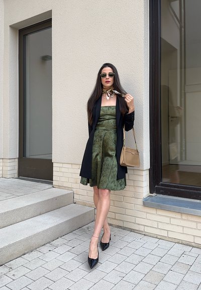 Vestido de satén verde con estampado floral, blazer negro, tacones negros de punta y un bolso pequeño beige. El cabello largo y unas gafas de sol completan el look.