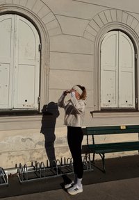 Une personne en veste blanche et leggings noirs se tient près d'un mur usé, ajustant ses lunettes de soleil tout en projetant une longue ombre. Banc vert à proximité.