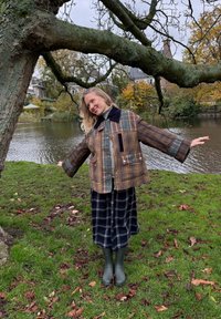 Brun rutete jakke med store lommer og en marineblå krage, båret over et svart rutete skjørt og grønne gummistøvler, stående ved en dam.