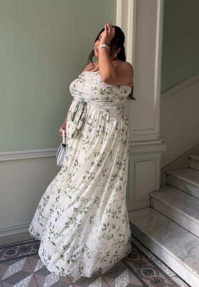 Vestido maxi blanco con estampado de flores y hojas verdes, diseño strapless, detalle de cintura anudada, falda fluida, combinado con un bolso plateado.