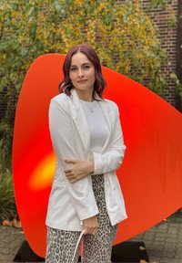 Bílý sako přes světlý top, spárované s kalhotami s leopardím vzorem. V pozadí přerostlá, lesklá oranžová socha.