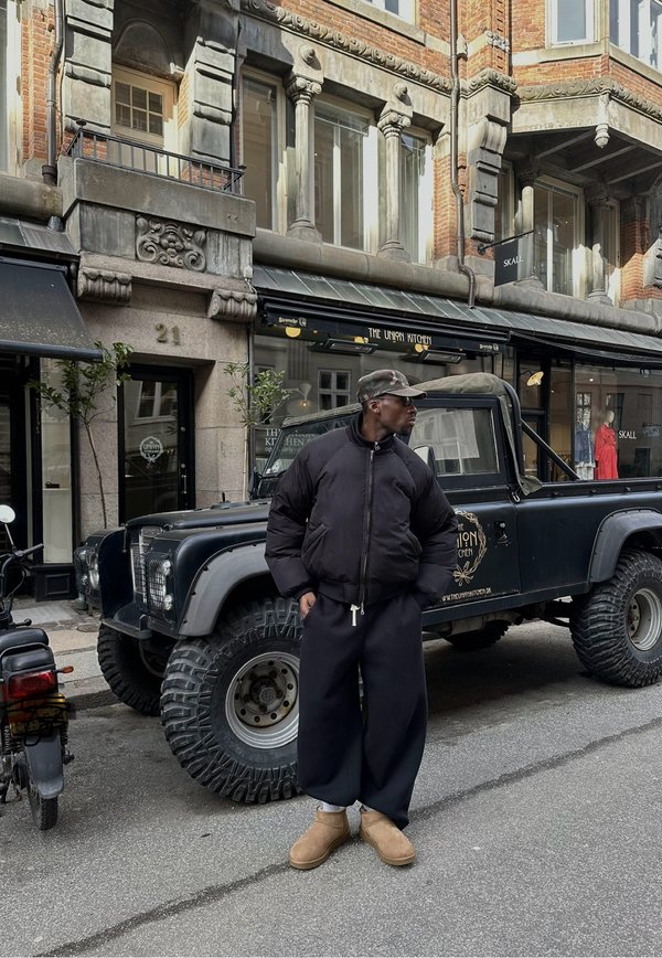 Homme portant une veste noire, une casquette, un pantalon large et des bottes marron clair, debout dans la rue à côté d'un camion tout-terrain noir avec le logo « The Union Kitchen ».