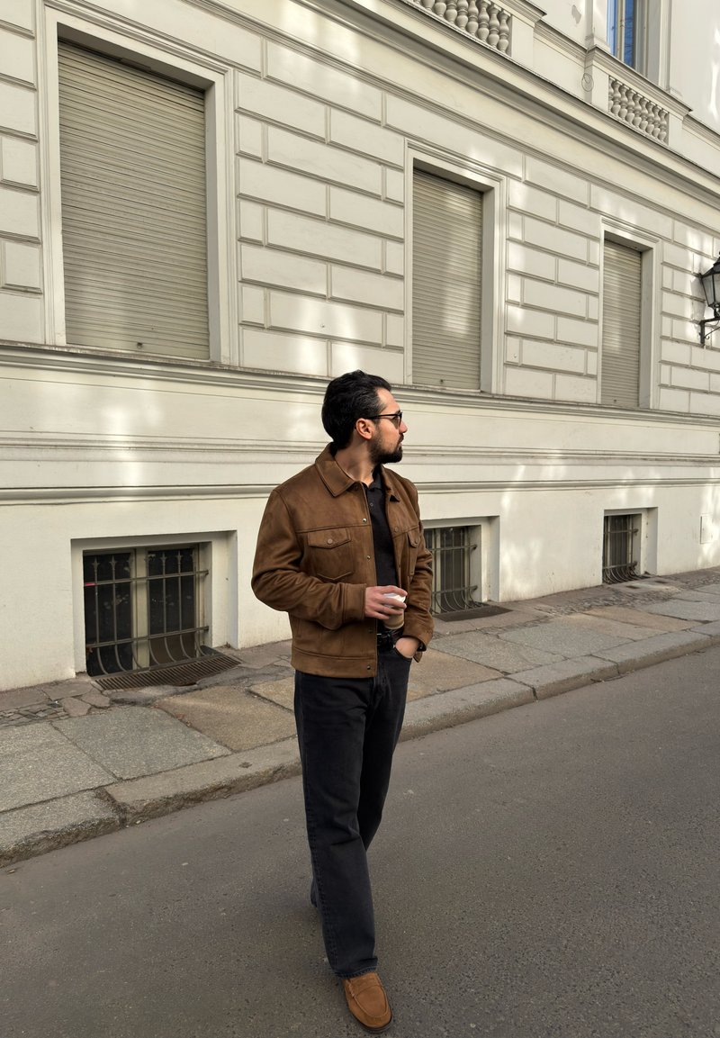 Homme en veste marron et pantalon noir tenant une tasse de café, debout dans une rue à côté d’un bâtiment blanc avec des fenêtres à volets fermés.