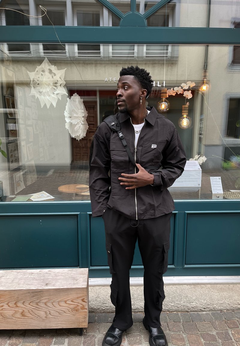 Veste noire à zip, à manches longues et avec des poches sur la poitrine, associée à un pantalon cargo assorti, le tout devant un décor illuminé.