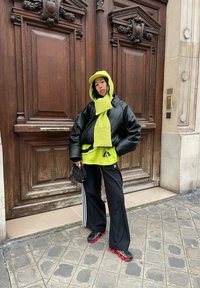 Črna oversize usnjena jakna, neon zelen hoodie in šal, črne atletske hlače z belimi črtami in črno-rdeči športni copati. Stoji ob lesenih vratih.