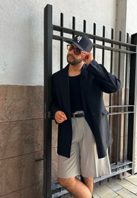 Černý sak na míru přes černou košili, doplněné světle šedými kraťasy. Ozdobeno černou čepicí a oversized brýlemi. Stojí blízko brány.