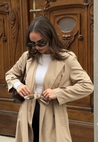 Beige trenchcoat med knytt liv, lange ermer og sidelommer; brukt over en hvit topp. Glatt materiale, supplert med svarte solbriller.