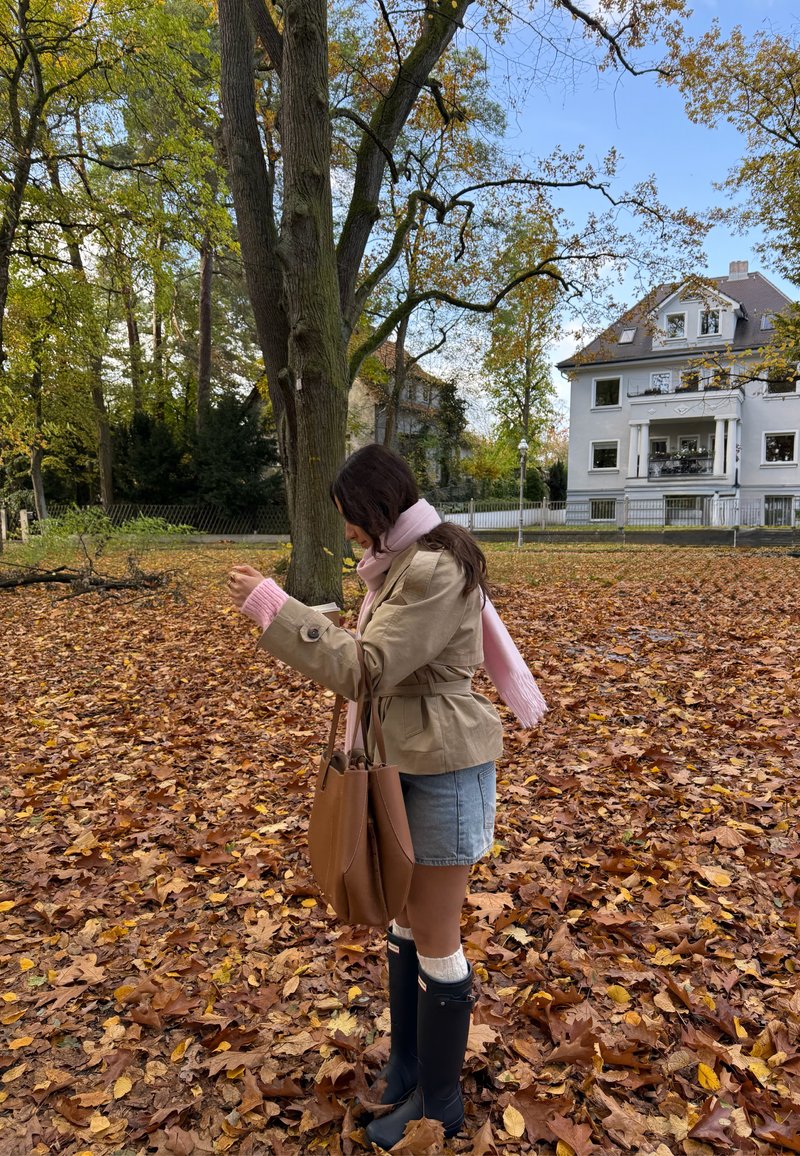 Trench coat castanho claro com cinto, usado sobre um cachecol cor-de-rosa. Combinado com uma saia de ganga, botas pretas até ao joelho e uma bolsa tote castanha na mão.