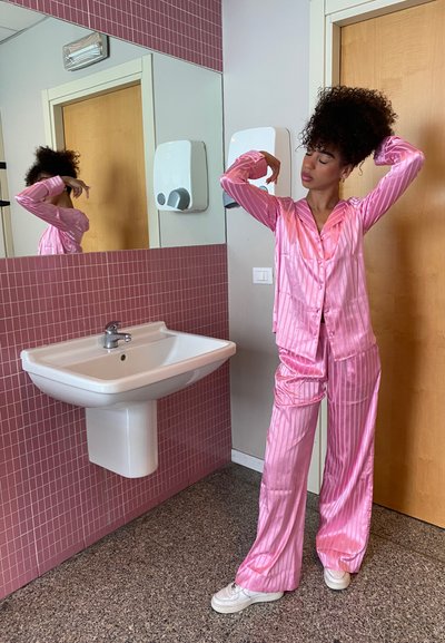Conjunto de lencería de satén rosa con rayas verticales, que incluye una blusa abotonada y pantalones holgados. En el fondo, se ven un lavabo y una pared con azulejos.