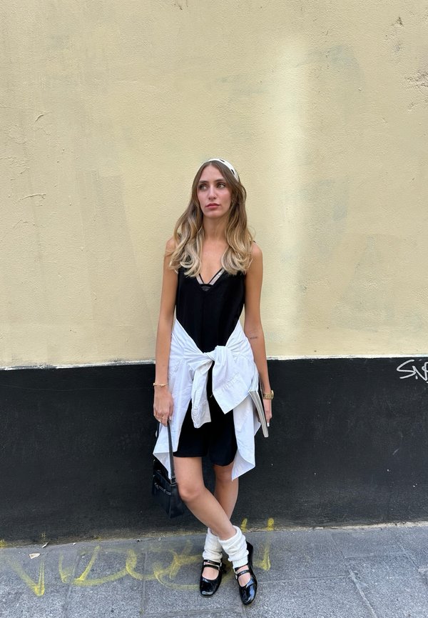 Joven mujer con el pelo largo está de pie junto a una pared beige y negra, lleva un vestido negro, camisa blanca anudada a la cintura, calentadores blancos para las piernas y zapatos negros.
