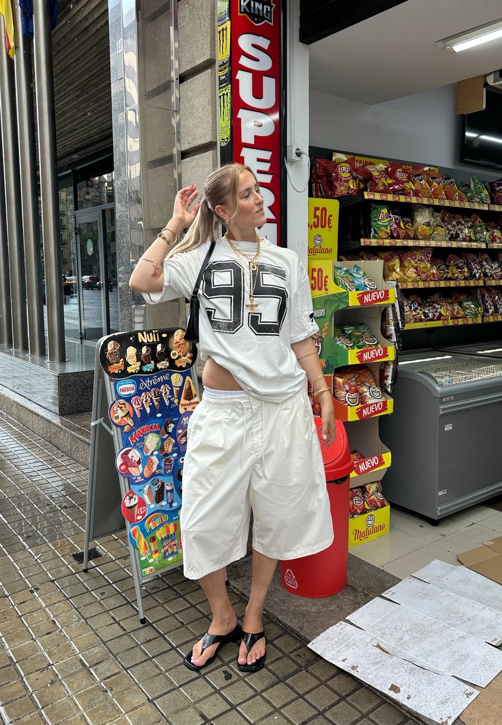 Fehér, over-size póló fekete "95" számmal, fehér, bő rövidnadrág, fekete flip-flop papucs és réteges arany nyakláncok. A háttérben egy kisbolt látható.