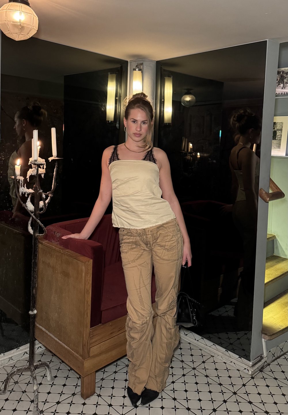 Femme appuyée contre un stand en bois dans une pièce faiblement éclairée avec un chandelier allumé, portant un haut beige, un pantalon à motifs et tenant un sac à main noir.