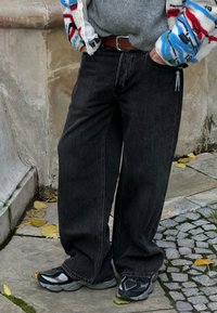 Jeans noirs à jambes larges avec une texture lisse, associés à des baskets noires et grises. La tenue est complétée par une couche extérieure colorée et à motifs.