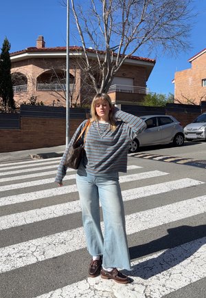 Joven con suéter a rayas, vaqueros azul claro y zapatos marrones de pie sobre un paso de peatones soleado en una zona residencial.