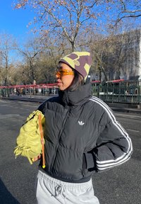 Personne portant une veste Adidas noire, un bonnet violet et jaune, des lunettes de soleil orange, tenant un sac jaune, debout dans une rue ensoleillée bordée d'arbres.