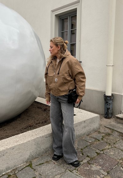 Chaqueta con capucha marrón con bolsillo delantero, pantalones anchos grises, bolso negro, de pie junto a una gran escultura esférica contra una pared neutra.