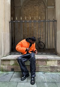 Veste à coussins orange avec une doublure noire, associée à un pantalon noir froissé et des bottes noires à semelle épaisse. Mur en briques texturé en arrière-plan.