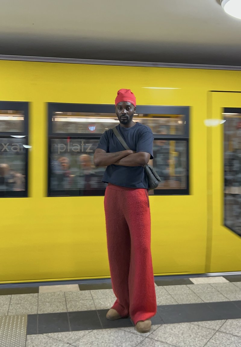 Pantaloni largi de culoare roșie, cu textură; tricou negru. Batic roșu pe cap. Persoana stă în fața unui tren galben cu ferestre care arată pasageri neclari.