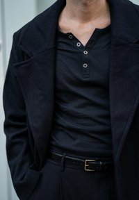 Černý vlněný kabát přes černé tričko henley s třemi knoflíky. Černý opasek se zlatou sponou drží černé kalhoty. Viditelné detaily z texturované tkaniny.