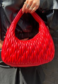 Bolsa de mão em couro sintético vermelho com design acolchoado, apresentando uma alça arredondada e textura suave. Acessórios em dourado são visíveis.