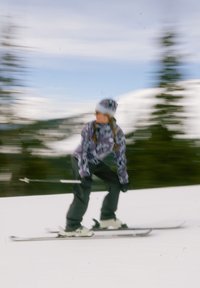 Persona esquiando cuesta abajo sobre la nieve, vistiendo una chaqueta con estampado, pantalones negros, gorro blanco y gafas, con un fondo de árboles en movimiento borroso.