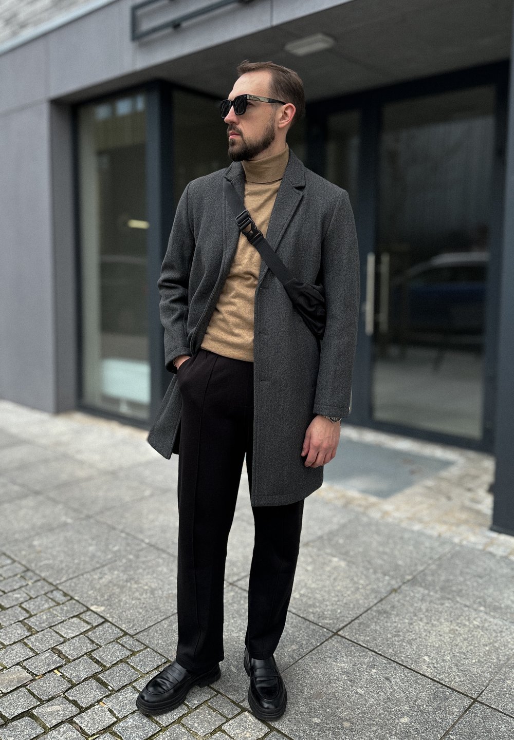Abrigo de lana gris sobre un suéter de cuello alto beige, pantalones negros y zapatos de cuero negros. Los accesorios incluyen gafas de sol oscuras y un bolso cruzado negro.