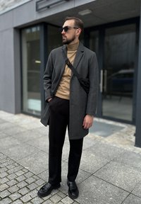 Abrigo de lana gris sobre un suéter de cuello alto beige, pantalones negros y zapatos de cuero negros. Los accesorios incluyen gafas de sol oscuras y un bolso cruzado negro.