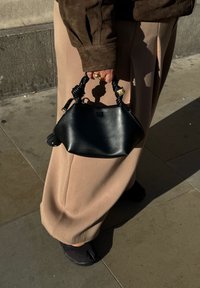 Bolsa de mão de couro preto com pega torcida preta, detalhes em hardware dourado e forma arredondada, segurada por uma pessoa vestindo calças bege.