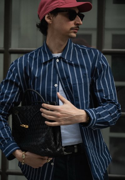 Camisa de botones a rayas en azul marino, bolso tejido negro, camiseta blanca, jeans oscuros y una gorra rosa. Las gafas de sol y la joyería completan el look.