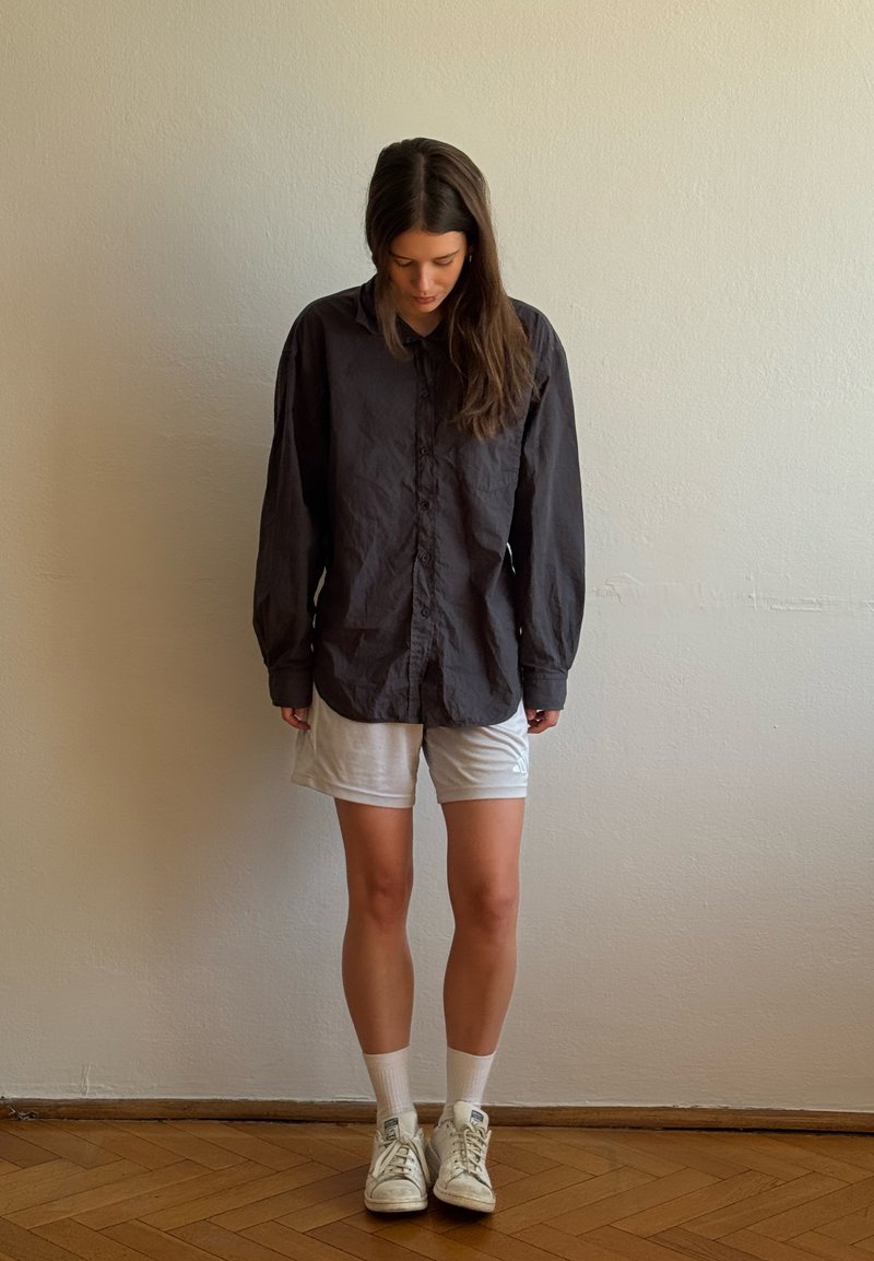 Femme aux longs cheveux portant une chemise noire oversize, un short blanc, des chaussettes blanches et des baskets blanches, debout sur un sol en bois et regardant vers le bas.