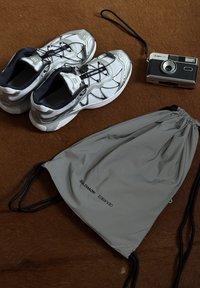 Ezüst atlétikai cipők texturált felsőrésszel, egy szürke logós húzózsinórs táska és egy retro fényképezőgép egy barna felületen.