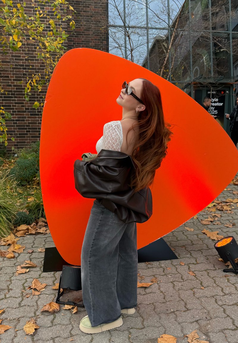 Veste marron en cuir surdimensionnée, haut en dentelle blanche, jean gris à jambes larges et chaussures vert clair, posant à côté d'un grand fond sculptural orange.