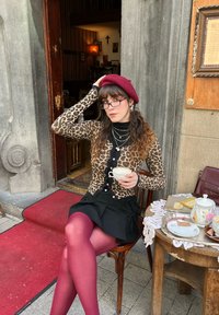Καρδιότι με λεοπάρ σχέδιο, μαύρη φούστα, μπορντώ μπερέ και ροζ καλσόν. Κρατώντας ένα φλιτζάνι τσαγιού, καθισμένο δίπλα σε ξύλινο τραπέζι με γλυκίσματα.