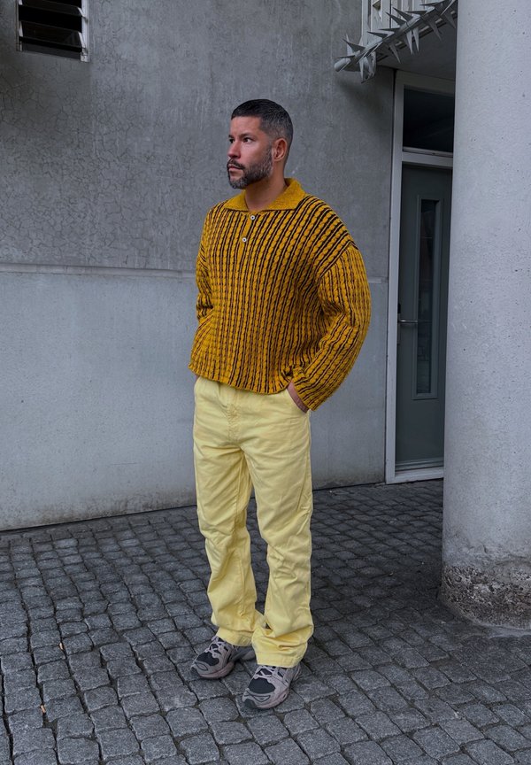 Maglione a righe gialle e nere, con collo e bottone, abbinato a pantaloni gialli chiari e sneakers grigie, in piedi su ciottoli.