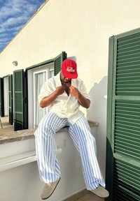 Man in a red cap, white shirt, and striped trousers is sitting on a ledge, adjusting his necklace outside a building with closed green shutters.