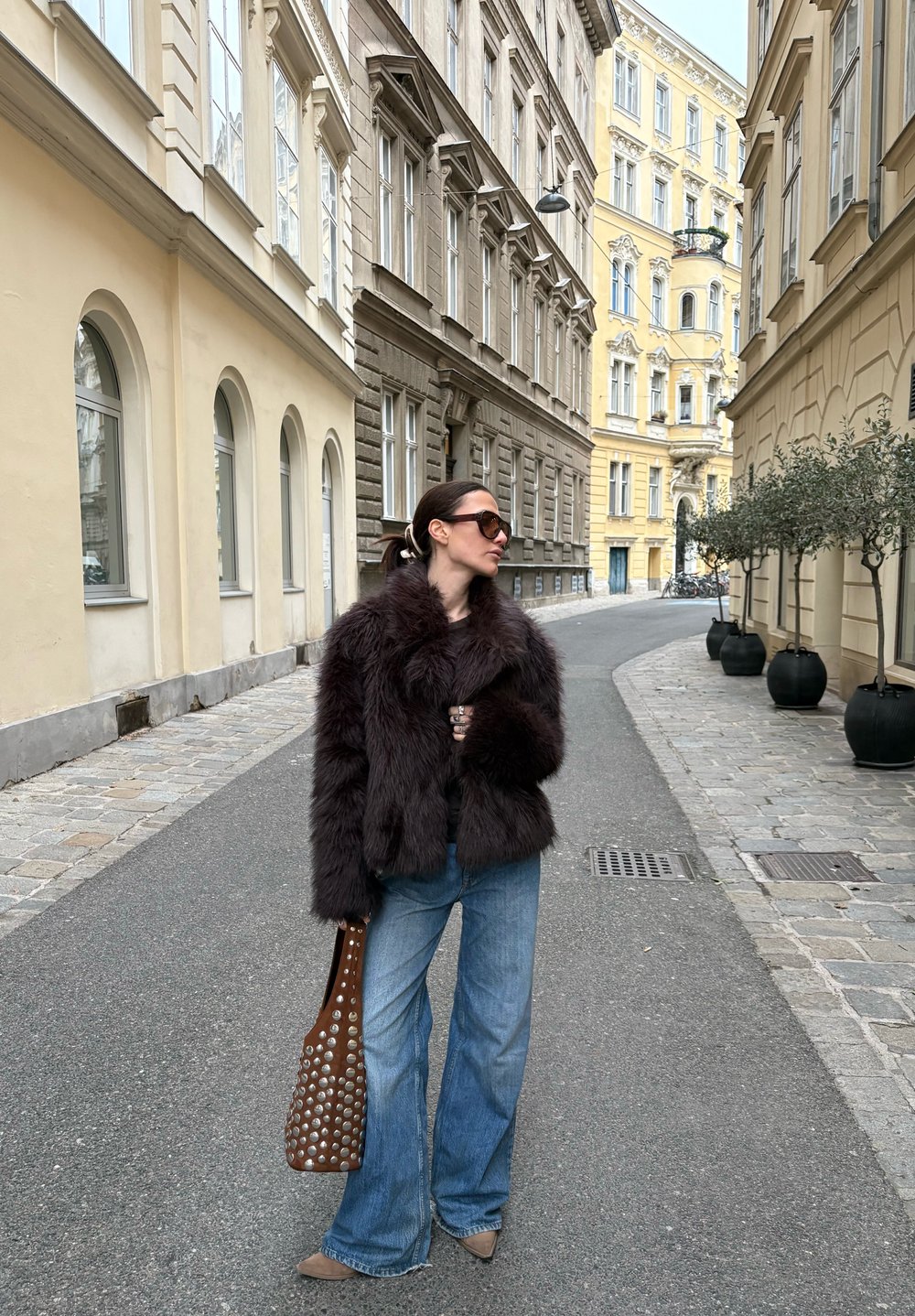 Veste en fausse fourrure bordeaux, jean évasé en denim, sac à main marron perforé. Rue pavée bordée de bâtiments aux tons doux.