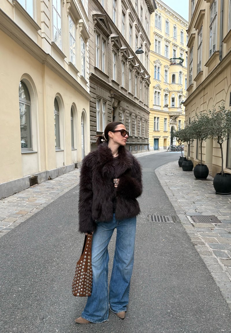Veste en fausse fourrure bordeaux, jeans évasés en denim, sac à main marron perforé. Rue pavée bordée de bâtiments aux tons doux.