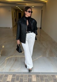 Sort læderjakke, sort rullekrave, hvide bukser med vide ben, sort håndtaske og sko med slangeprint, i et moderne interiør.