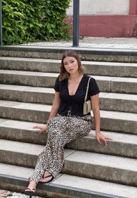 Nero top a maniche corte con nodo frontale, abbinato a una gonna con stampa leopardata. Sandali neri opachi. Borsa a tracolla chiara. Scale di pietra.