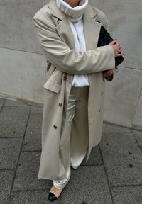 Femme en trench-coat beige et pantalon en satin, pull col roulé blanc, tenant une pochette noire, debout sur un trottoir carrelé.